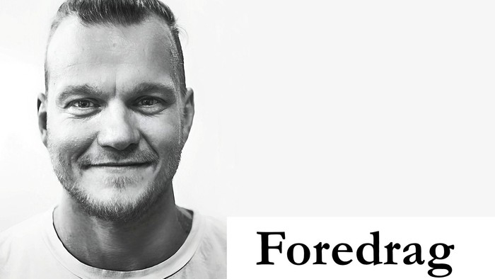 Sort-hvidt portræt af en smilende mand med kort hår og skæg, mærket **