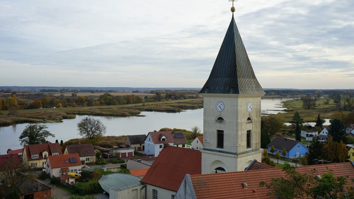 Kirchturm mit spitzem Dach, umgeben von Häusern mit roten Dächern, Flusslandschaft im Hintergrund