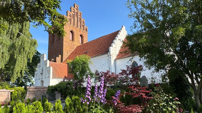En charmerende historisk kirke omgivet af frodig grønne områder og farverige blomstrende planter under en klar himmel.