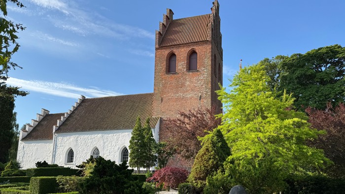 Historisk kirke med rødt murstens tårn omgivet af omhyggeligt anlagt grønne områder under en klar himmel.