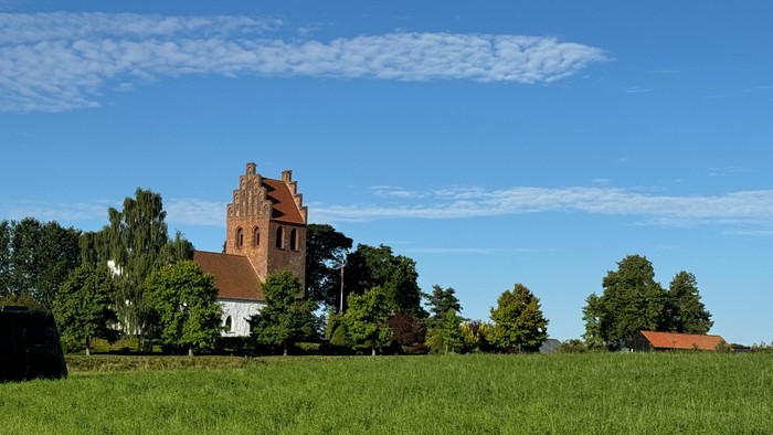 En malerisk kirke med rødt tag står midt i frodige grønne marker under en klar blå himmel.