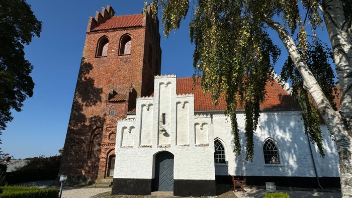 Et gammelt teglsteinskirke med tårn og hvide detaljer, omgivet af græs og træer