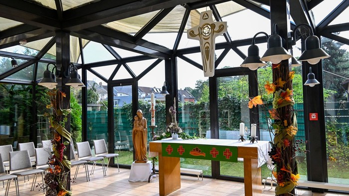 Moderne Kapelle mit Glaswänden, die einen ruhigen Altar zeigt, der mit Blumenschmuck und religiösen Statuen verziert ist.