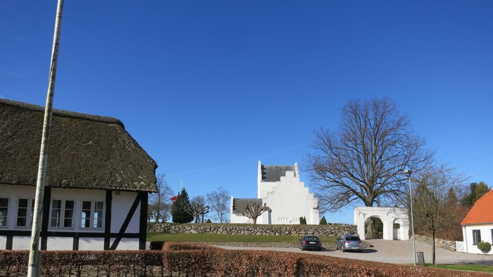 Hvid bygning med bil foran, omgivet af grønne omgivelser og klar blå himmel.