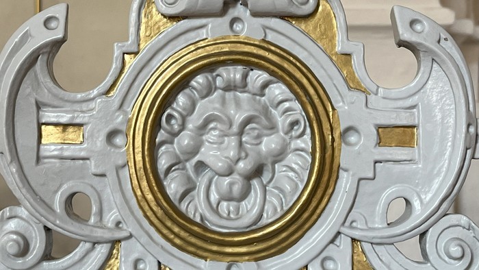 Udsmykket dekorativ plade med en løvemaskeramme omgivet af indviklede guld- og hvide detaljer.