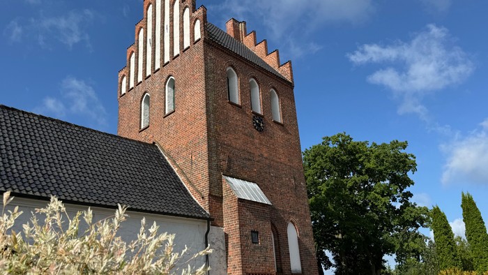 Historisk murstenskirketårn med stejlt gavltag og buede vinduer omgivet af grønne områder.
