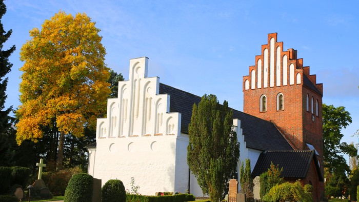 En moderne hvid bygning og en rød kirke med tårn ved en park med høstfarvede træer
