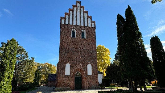 Historisk murstenskirketårn med buet indgang, omgivet af velplejet beplantning og træer.