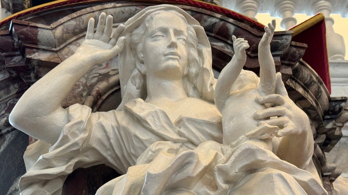 En marmorstatue af en liggende kvinde med udstrakte arme, sandsynligvis af religiøs karakter, placeret mod en marmorbaggrund.