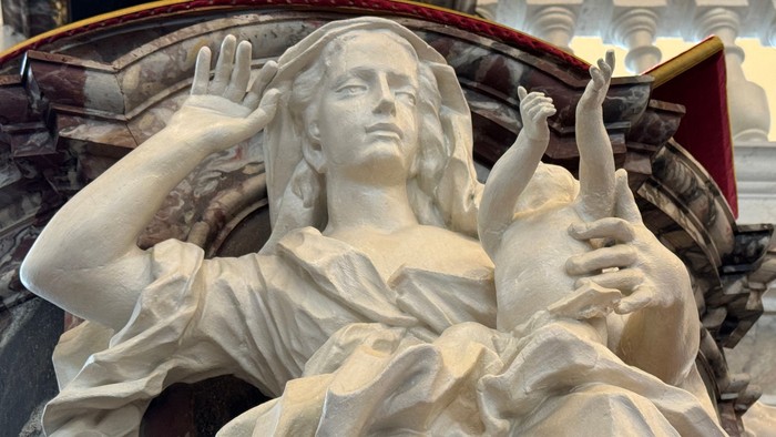 En marmorstatue af en liggende kvinde med udstrakte arme, sandsynligvis af religiøs karakter, placeret mod en marmorbaggrund.