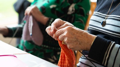 En person spinder med en orange garn, mens en anden person sitter i baggrunden.