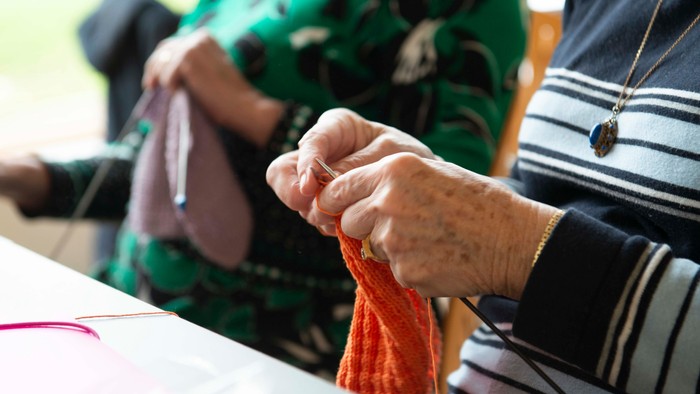 En person spinder med en orange garn, mens en anden person sitter i baggrunden.