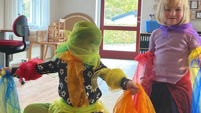 To børn i farverige kostumer leger med farvestrålende stof indendørs og ser ud til at danse eller spille skuespil.