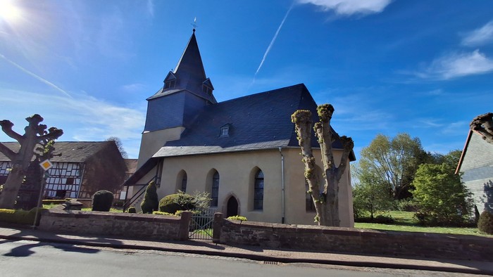 Kirche mit hohem Turmspitz und Kreuz, umgeben von Grünflächen unter blauem Himmel.