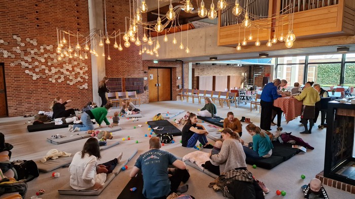 En stor hall med mange mennesker, der sidder på gulvmatte og deltager i aktiviteter under hængende lys.