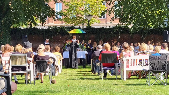 En gruppe af mennesker sidder på stolene i en have og lytter til en person, der taler ved et bord med en grøn paraply.