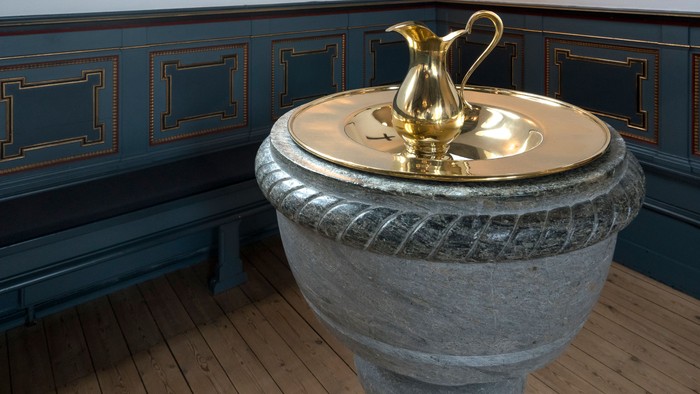 Antikt messingkande oven på en sten døbefont i et træindrettet interiør.