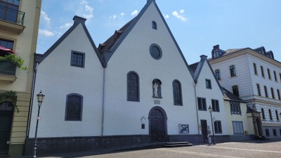Weißes Kirchengebäude mit schwarzem Dach und Fenstern, umgeben von Gebäuden auf Kopfsteinpflasterplatz
