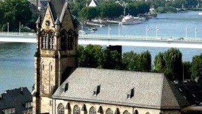 Eine Kirche mit hohem Turm steht neben einem Fluss, im Hintergrund eine Stadtlandschaft.