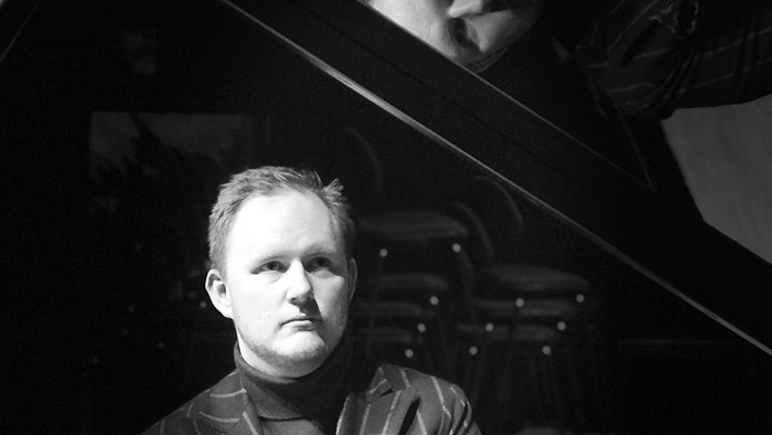 En mand i formelt tøj sidder eftertænksomt ved siden af et stort flygel i et svagt oplyst rum.