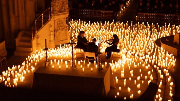Two people sit among numerous lit candles in a dimly lit area.