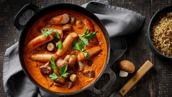 Hjertelig gryderet med pølser, svampe og urter serveret i en rustik gryde ved siden af friske ingredienser.