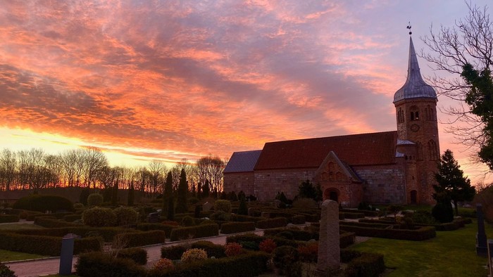 En kirke med tårn og klokke i solnedgang med rød og orange himmel, omgivet af en gravplads og grønne planter.