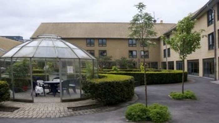 En lille glasspavillon i en courtyard med grønne planter og et hus i baggrunden