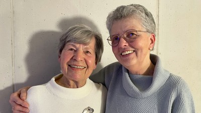 Zwei ältere Frauen lächeln und stehen eng beieinander, die Arme umeinandergelegt.