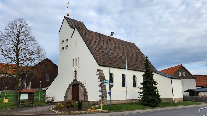Kleine weiße Kirche mit spitzem Dach und Kreuz auf dem Turm, umgeben von Bäumen und Häusern.