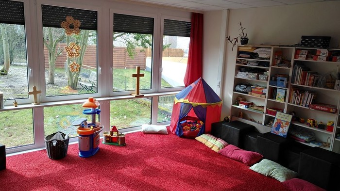 Kinderzimmer mit Spielsachen und Büchern, große Fenster, roter Teppich.