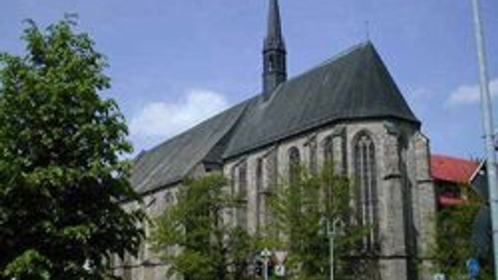 Große Kirche mit hohem Turm und spitzen Dach, umgeben von Bäumen und einem Auto.