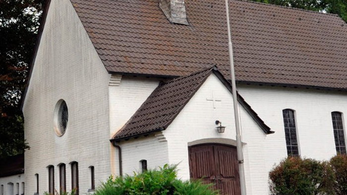 Weiße Kirche mit hohem Turm und braunem Dach, umgeben von Bäumen und Büschen