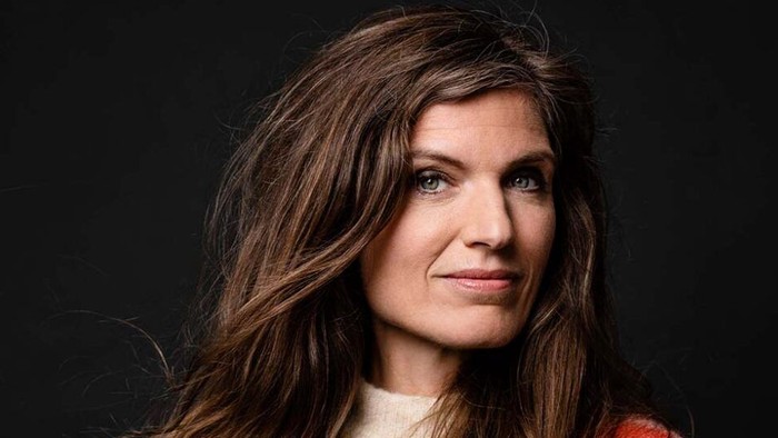 En kvinde med langt, bølget brunt hår krydser armene i en hyggelig, stribed trøje mod en mørk baggrund.