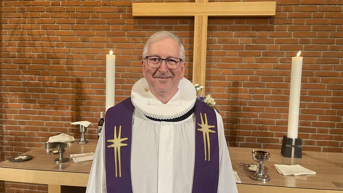 En gejstlig i liturgisk messeklædning står foran et kors i en kirkelig omgivelse med lys og altergenstande.