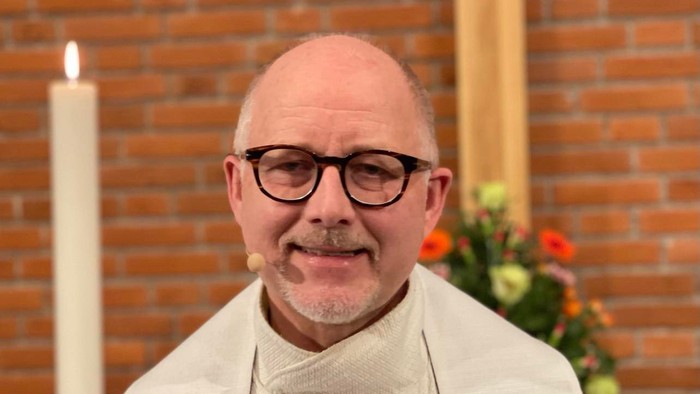 En bald, brillet mand i hvid liturgisk klæde foran mur med lysende lys og blomster.