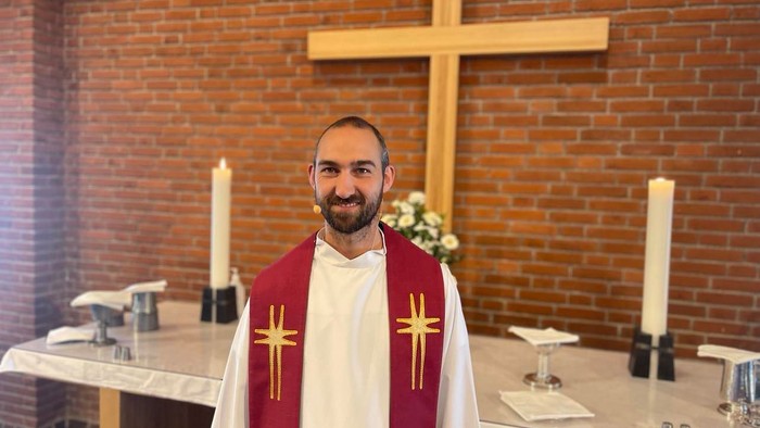 Mand i rødt og hvide præsteklæder foran alter med kors og lysstake