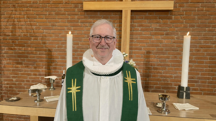 En mand i gejstligt tøj står i en kirke iført briller og en hvid krave med kors.