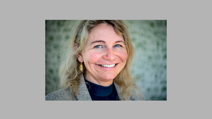 Kvinde med smilende ansigt, blondt hår iført en grå jakke og mørkt top på en neutral baggrund.