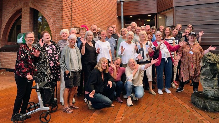 En glad gruppe mennesker i forskellige aldre, der poser sammen udendørs med festlige håndbevægelser.