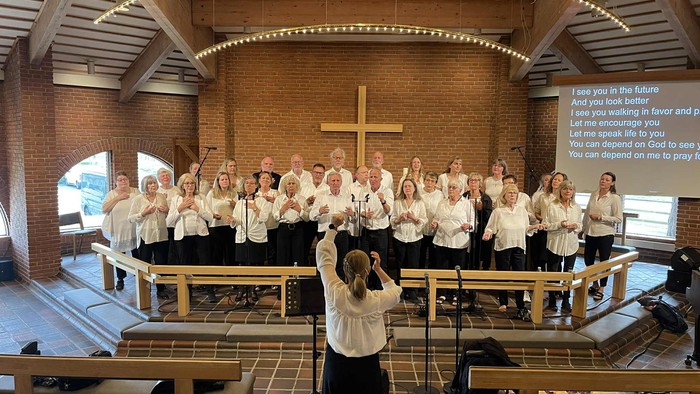 Et kor optræder i en kirke, ledet af en dirigent med et stort kors og opbyggelige skriftsteder udstillet.
