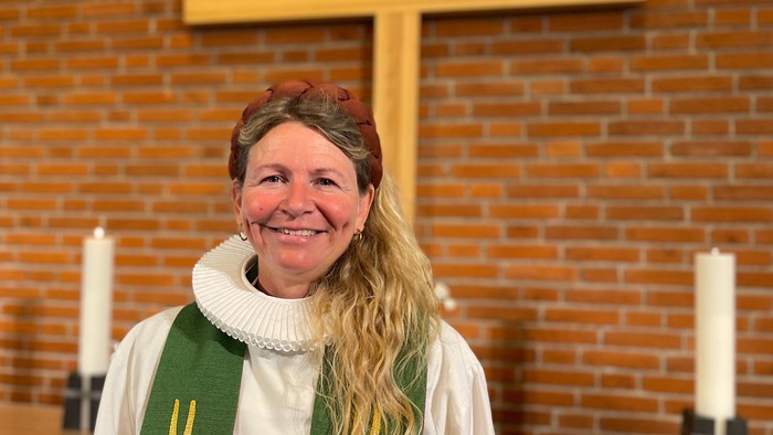 En person i religiøst tøj står og smiler i en kirke med et stort kors og lys bag sig.