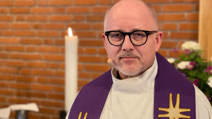 En baldrigt skaldet mand i religiøse klæder foran mursten og lysende stearin.