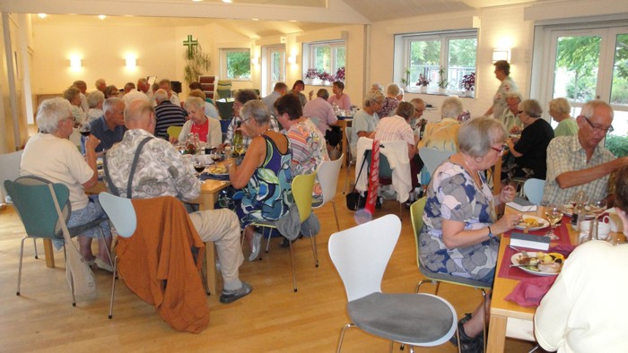 En gruppe ældre folk sidder og spiser sammen i en lys og rummelig lokalitet.