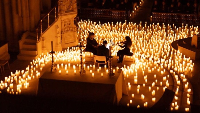 People sit in solemn silence among rows of lit candles in a dimly lit cathedral.