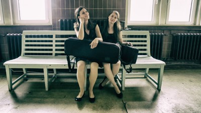 Zwei Frauen sitzen auf getrennten Bänken, beide in Schwarz gekleidet, wobei die eine den Kopf hält und die andere zur Seite blickt.