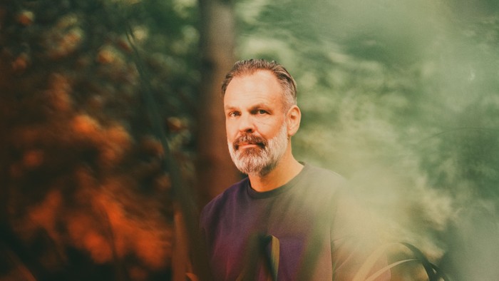 En mand med skæg står i en skov, hvor sollyset filtrerer ned mellem træerne.