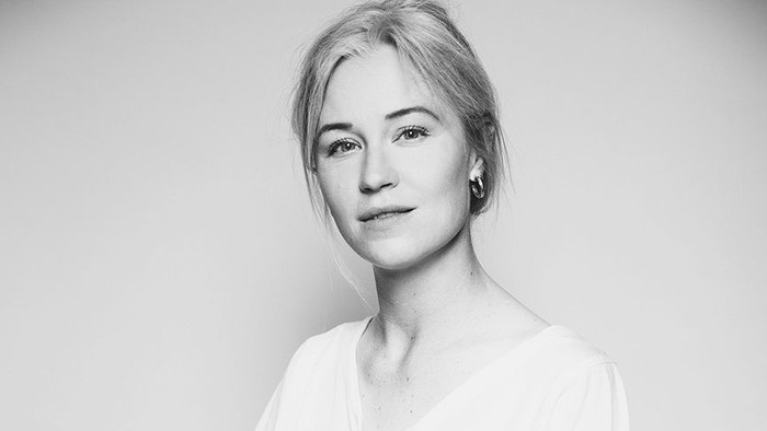 En kvinde i en hvid bluse og sorte bukser sidder elegant på en skammel i et minimalistisk sort-hvidt portræt.