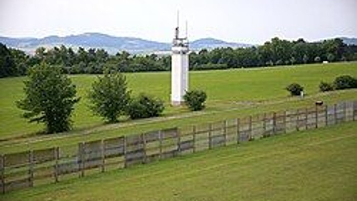 Ein hoher weißer Leuchtturm steht auf einem grasbewachsenen, eingezäunten Feld mit fernen Hügeln und Bäumen.