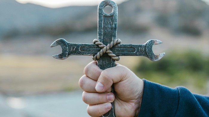 Eine Hand hält eine kreuzförmige Schraubenschlüssel-Anordnung vor einem malerischen Outdoor-Hintergrund.